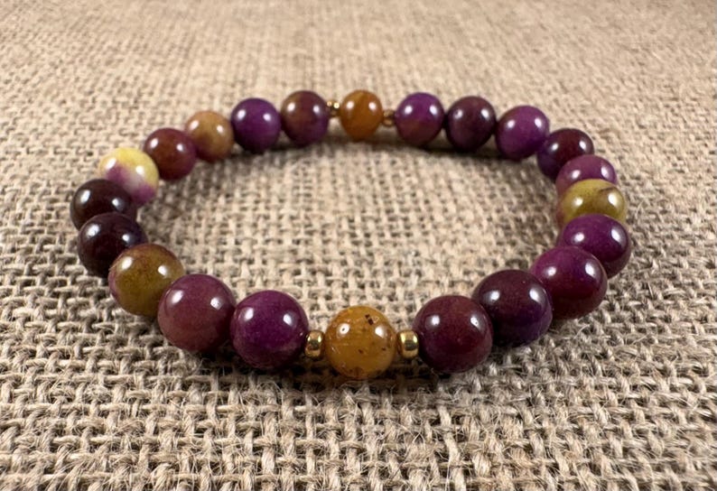 May include: A beaded bracelet featuring smooth, round beads in shades of deep purple, magenta, and mustard yellow. The beads are arranged in a circular pattern and are accented by small, polished gold-toned metal spacers placed between the beads.