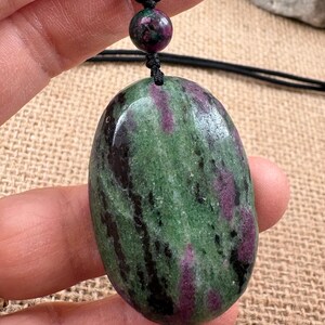 May include: Oval-shaped pendant necklace featuring a polished stone with green, purple, and black marbling. The pendant is suspended from a black cord with a matching bead. The stone's natural colors and patterns create a unique, earthy aesthetic.