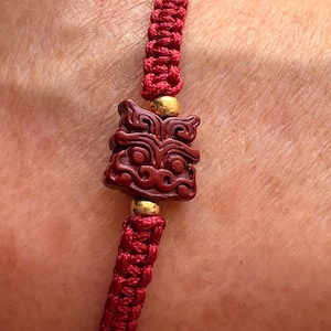 May include: Red braided bracelet featuring a carved dark red centerpiece with a detailed face design. The bracelet is accented with gold-colored beads. The braided cord is a vibrant red color.