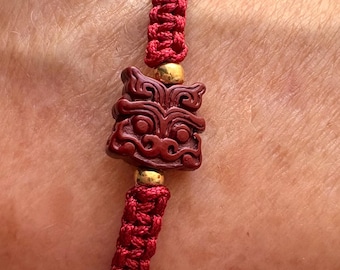 May include: A red braided cord bracelet featuring a central dark red carved rectangular charm with a stylized face design, flanked by two small metallic gold-colored spherical beads, secured with a woven knot pattern along the entire band.
