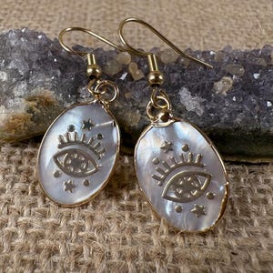 May include: Gold-tone earrings with a pearlescent oval pendant. Each earring features an eye and star design. The earrings are hanging on a gold-colored hook.