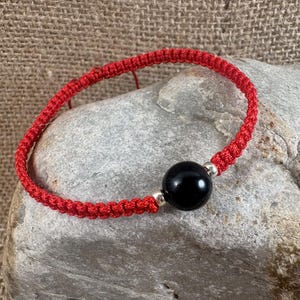 May include: Red macrame bracelet with a large, round, black bead and silver-colored accents. The bracelet is knotted with a bright red cord and is resting on a gray stone surface. The bracelet is a simple, yet elegant accessory.