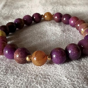 May include: A stretch bracelet composed of round, polished beads in shades of deep purple and mottled yellow-orange. The beads are arranged in a circular pattern, interspersed with small, metallic gold-toned spacer beads between the larger yellow-orange beads.