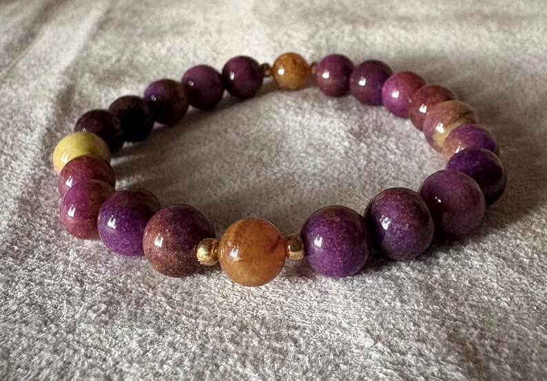 May include: A stretch bracelet composed of round, polished beads in shades of deep purple and mottled yellow-orange. The beads are arranged in a circular pattern, interspersed with small, metallic gold-toned spacer beads between the larger yellow-orange beads.