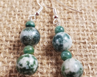 May include: A pair of dangle earrings featuring a silver hook and three beads in shades of green and white. The earrings have a natural, organic look, with the beads varying in size and pattern. The background is a textured, neutral-toned surface.
