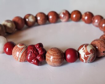 May include: A stretch bracelet composed of round, polished beads in shades of deep red, terracotta, and cream. The beads feature natural swirling patterns and textures. A single carved bead shaped like a cat paw is centered among the stones.