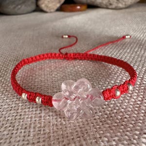 May include: Red macrame bracelet featuring a clear, flower-shaped crystal centerpiece and small silver beads. The adjustable bracelet is displayed on a textured, neutral-colored surface. A delicate and feminine accessory.