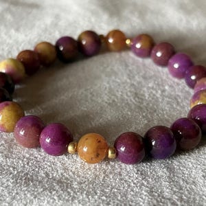 May include: A stretch bracelet composed of round, polished beads in varying shades of deep purple, magenta, and golden yellow. The beads are arranged in a circular pattern and are accented by small, smooth, metallic gold-toned spacer beads.