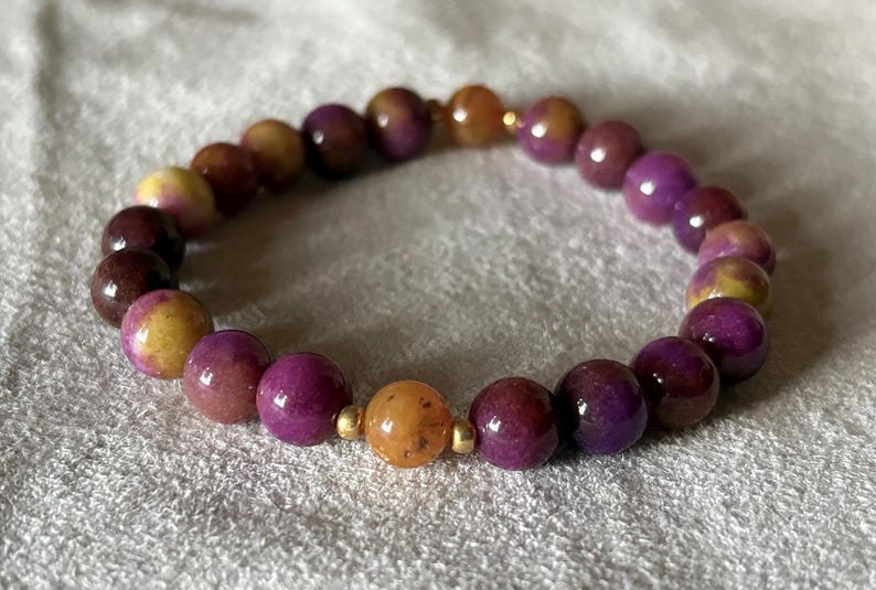 May include: A stretch bracelet composed of round, polished beads in varying shades of deep purple, magenta, and golden yellow. The beads are arranged in a circular pattern and are accented by small, smooth, metallic gold-toned spacer beads.