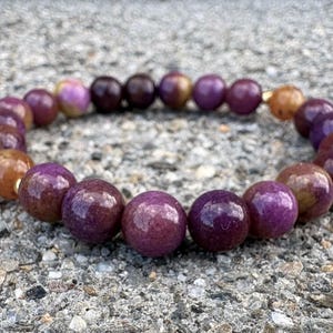 May include: A circular beaded bracelet featuring polished, spherical stones in varying shades of deep purple, magenta, and warm amber-brown. The beads are strung together in a continuous loop, showcasing a smooth, glossy finish and natural color variations.