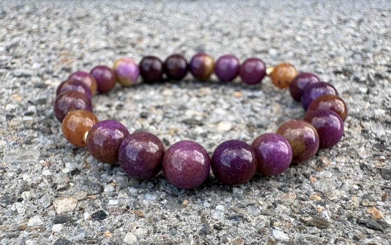 May include: A circular beaded bracelet featuring polished, spherical stones in varying shades of deep purple, magenta, and warm amber-brown. The beads are strung together in a continuous loop, showcasing a smooth, glossy finish and natural color variations.