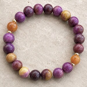 May include: A circular beaded bracelet featuring smooth, round stones in shades of deep purple, magenta, and golden yellow. The beads are arranged in a repeating pattern and are separated by small, polished gold-tone metal spacer beads.