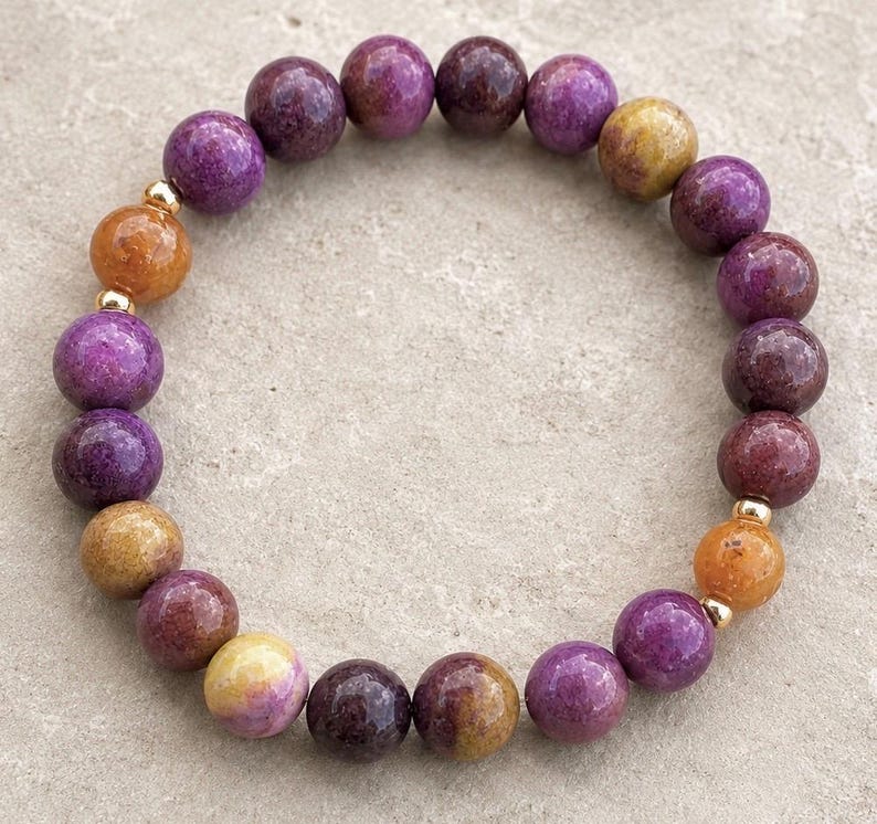 May include: A circular beaded bracelet featuring smooth, round stones in shades of deep purple, magenta, and golden yellow. The beads are arranged in a repeating pattern and are separated by small, polished gold-tone metal spacer beads.