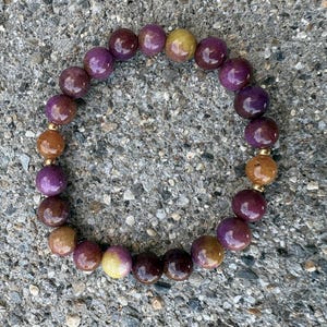 May include: A circular beaded bracelet featuring polished, spherical beads in shades of deep purple, plum, and warm amber. The bracelet is strung with small, metallic gold-toned spacer beads placed between the larger, smooth, multi-colored stones.