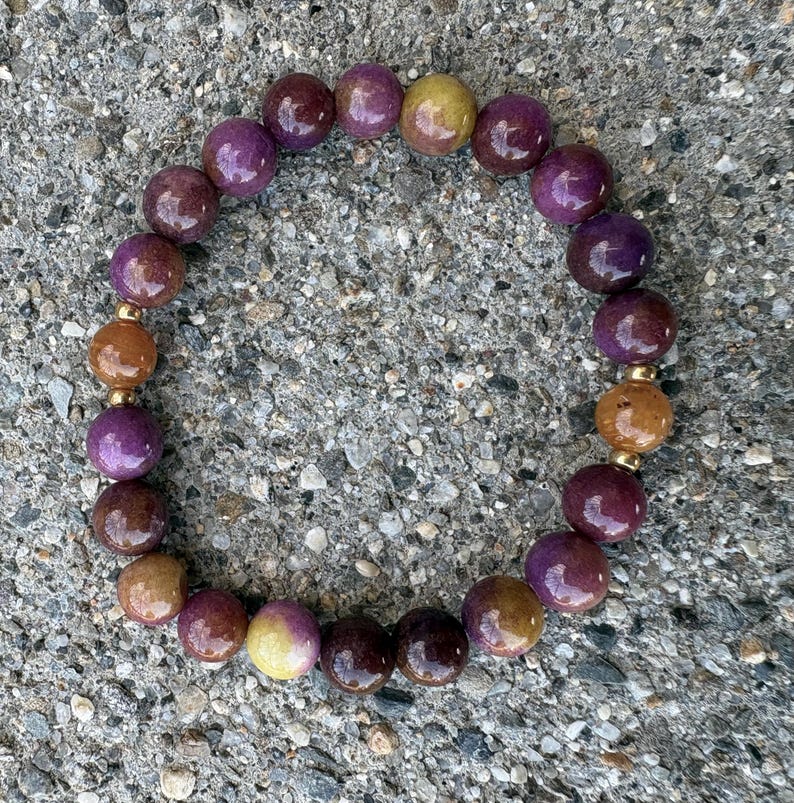 May include: A circular beaded bracelet featuring polished, spherical beads in shades of deep purple, plum, and warm amber. The bracelet is strung with small, metallic gold-toned spacer beads placed between the larger, smooth, multi-colored stones.