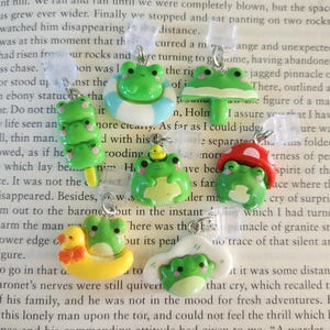 Könnte beinhalten: Eine Sammlung grüner Frosch-Anhänger mit verschiedenen Designs, darunter ein Pilz, ein Eis am Stiel, eine Ente und eine Schwimmweste. Jeder Anhänger hat eine silberfarbene Schlaufe zum Befestigen an Schmuck oder anderen Gegenständen.