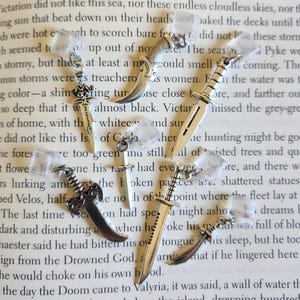 May include: A collection of small, silver-colored sword and dagger charms. The charms vary in design, with some featuring ornate handles and detailed blades. They are displayed on a book page.