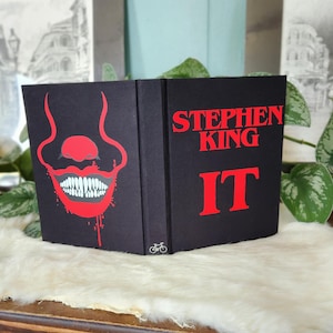 以下が含まれることがあります： 表紙が黒色のハードカバーの本で、赤いイラストが描かれています。イラストは、クレーンの口と歯です。本のタイトルは「IT」で、赤い文字で書かれています。作者の名前「STEPHEN KING」は、タイトルの上に白い文字で書かれています。