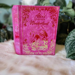 Puede incluir: Un libro rosa con detalles dorados y un título que dice "Alice's Adventures in Wonderland". La portada presenta una ilustración de Alicia cayendo por un agujero de conejo.