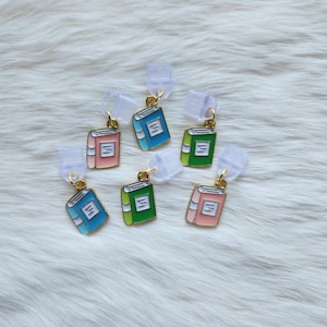 May include: Seven colorful book-shaped charms with gold accents. Each charm features a different color and a simple design of a closed book with a white spine.
