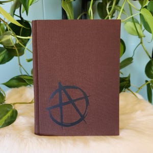 May include: A brown hardcover book featuring a black anarchy symbol printed on the front cover. The book has a textured, matte finish and is positioned vertically, displaying a clean, solid-colored surface with a distinct graphic icon.