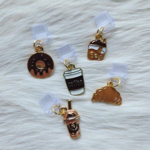 Peut inclure: Cinq breloques dorées avec différents motifs de nourriture et de boissons. Les breloques sont attachées à des clips en plastique transparent. Les motifs comprennent un beignet, une tasse de café, un thé à bulles, un croissant et un milk-shake.