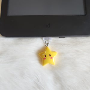 Puede incluir: Un dije amarillo en forma de estrella con una cara sonriente y mejillas rosadas cuelga de un dispositivo electrónico negro. El dije está sujeto con un anillo de metal plateado y un adhesivo de plástico transparente.