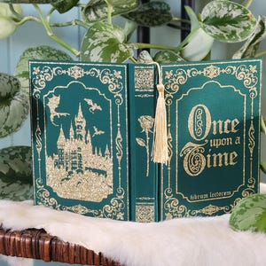 May include: A green book with gold glitter accents and the words "Once upon a Time" on the cover. The left side features a castle silhouette with bats. A gold tassel bookmark hangs from the spine. The book is open.