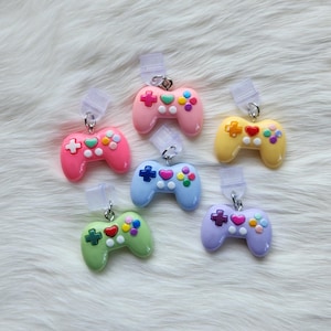 Puede incluir: Seis charms de mando de juego de plástico en colores pastel con botones coloridos. Los charms están unidos a cordones de plástico transparentes.