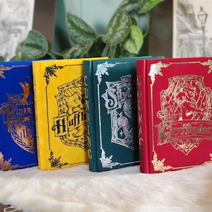 Könnte beinhalten: Vier Hardcover-Bücher mit verschiedenen farbigen Einbänden und goldgeprägten Wappen. Die Bücher sind betitelt mit "Ravenclaw", "Hufflepuff", "Slytherin" und "Gryffindor".