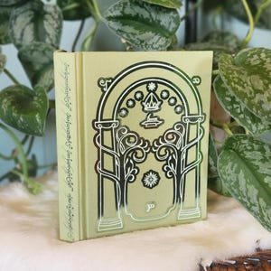 May include: A hardcover book with a light green cover and silver foil accents. The cover features an arched design with floral and geometric elements. The book is resting on a white, fluffy surface, with green foliage in the background.