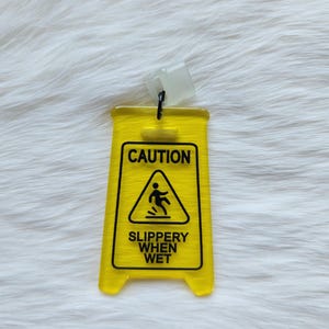 Puede incluir: Señal de precaución de plástico amarillo con texto negro que dice "CAUTION", una silueta negra de una persona resbalando y "SLIPPERY WHEN WET".
