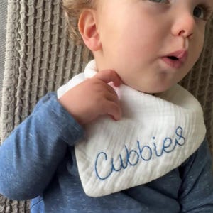 May include: White baby bib with the word "Cubbies" embroidered in blue script. The bib is made of a soft, textured fabric and is worn by a baby wearing a blue long-sleeved shirt.