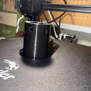 May include: A black 3D printed cylindrical object with a flat base and two small circular holes near the top rim. The object is positioned on a textured black print bed surface directly beneath the print head of a 3D printer machine.