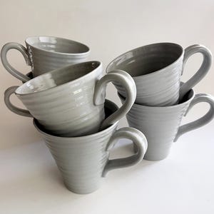May include: A collection of five light gray ceramic mugs, each featuring a tapered shape and a series of horizontal ridges along the exterior surface. Each mug includes a curved handle attached to the side, with a smooth, glossy finish.