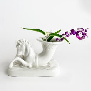May include: A white ceramic vase shaped like a horse with a flared horn-like opening on its back. The horse is depicted in a reclining position on a rectangular base. A single stem with green leaves and small purple flowers is placed inside.
