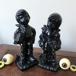 May include: Two black ceramic figurines of a boy and a girl. The boy is wearing overalls and holding a hat. The girl is wearing a dress and holding a book. Two realistic-looking eyeballs are next to the figurines.