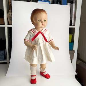 May include: A vintage doll with a cream-colored dress featuring a red bow and trim. The doll has short brown hair, green eyes, and is wearing red Mary Jane shoes and white socks. The doll is standing in front of a white backdrop.