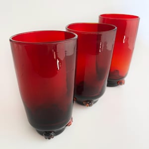 May include: Three red-tinted drinking glasses. The glasses have a cylindrical shape with a slightly flared rim. The base of each glass has a decorative, dark-colored design. The glasses are arranged in a row against a white background.