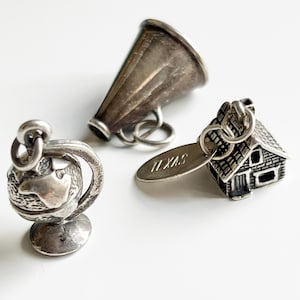May include: Three silver charms: a globe on a stand, a megaphone, and a house with a Texas tag. The globe charm features detailed continents. The megaphone has a classic shape. The house charm has a detailed roof and windows.