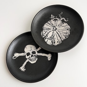 May include: Two black ceramic plates with white illustrations. One plate features a skull and crossbones, the other a pumpkin with a butterfly and bee. The plates are likely for Halloween or themed dining.