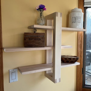 May include: A white wooden shelf unit with four shelves. The shelves are stacked in a staggered pattern. A small wooden box with the word "RECIPES" on it is on the second shelf. A white ceramic jar with the word "COFFEE" on it is on the top shelf. A vase with purple flowers is on the first shelf.