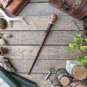 Op de afbeelding: Een donkerbruine houten toverstaf met een decoratieve, gespikkelde, lichtbruine bol aan de bovenkant. De toverstaf is omgeven door antieke sleutels, leren koffers en potten met kurken deksels op een verweerd houten oppervlak.