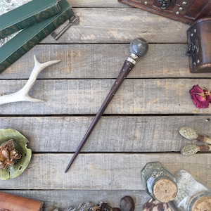 May include: A dark wooden wand with a gray marble sphere at the top and silver accents. The wand is long and slender, with small silver studs along the shaft. The background includes books, a key, and other decorative items.