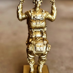 Puede incluir: Una figurita dorada de un paracaidista con un paquete de paracaídas y un casco. La figurita está de pie sobre una base dorada.