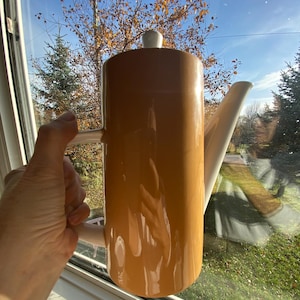 May include: A tall, mustard-yellow ceramic coffee pot with a white spout, handle, and lid. The pot is held in front of a window, reflecting the outdoor scenery. The pot has a glossy finish and a cylindrical shape.