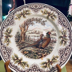 May include: A round ceramic plate featuring a central illustration of two pheasants in a landscape with trees and a fence. The rim is decorated with a repeating brown and green pattern of oak leaves and acorns on a cream-coloured background.