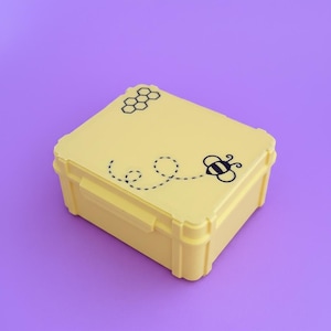 May include: A yellow rectangular box with a black bee and honeycomb design. The bee is depicted with a dotted flight path. The box has a secure latch and is set against a purple background.
