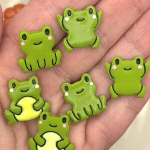 May include: Six small, green frog-shaped charms with black eyes, pink cheeks, and white accents. Some frogs feature a light yellow belly, while others are solid green. Each frog has a smooth, flat surface with detailed black outlines.