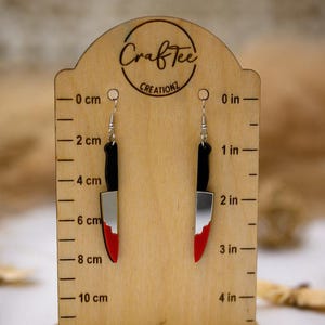 Peut inclure: Une paire de boucles d'oreilles en forme de couteau argentées avec des accents rouges. Les boucles d'oreilles sont présentées sur un support en bois avec une règle en centimètres et en pouces.